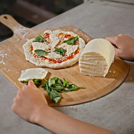  Eine Person, die eine Pizza auf einer hölzernen Pizzaschaufel mit frischem Mozzarella, Tomatensoße und Basilikum vorbereitet. Ein großer Käseblock und frische Basilikumblätter liegen daneben.